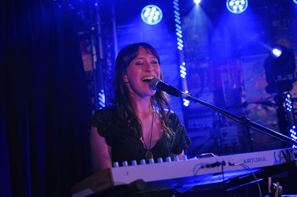 Female singer with eyes closed, singing into a microphone while playing a white keyboard on a blue-lit stage.